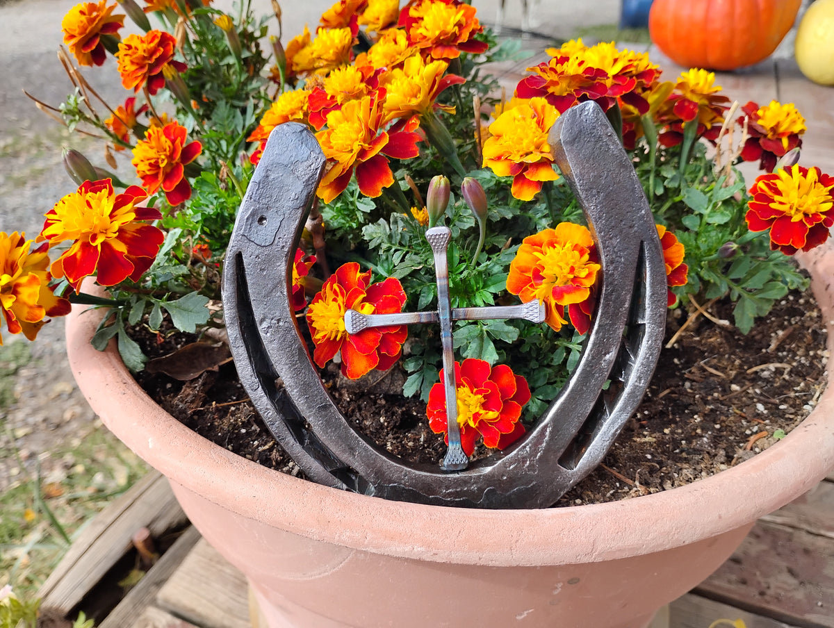 Handmade Christian horseshoe nail cross displayed in a terra cotta pot of orange and red marigolds — a single horseshoe with a cross made from horseshoe nails centered inside, handcrafted Western faith décor