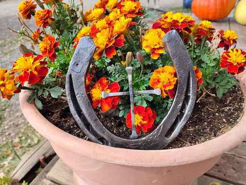 Handmade Christian horseshoe nail cross displayed in a terra cotta pot of orange and red marigolds — a single horseshoe with a cross made from horseshoe nails centered inside, handcrafted Western faith décor