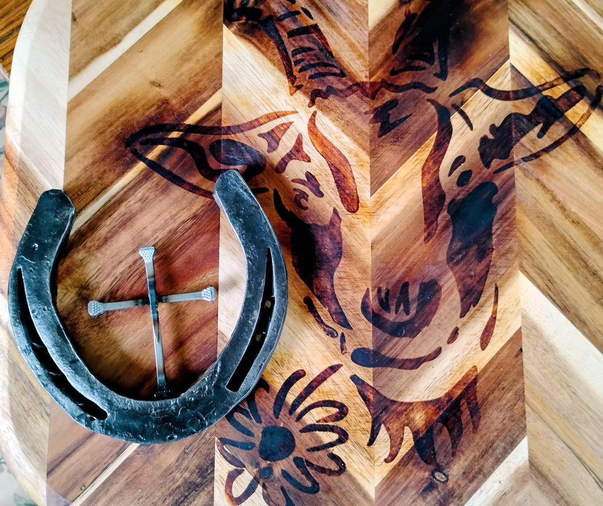 Handmade horseshoe nail cross inside a single horseshoe halo, displayed on a decorative wood-burned chevron cutting board with Western designs — handcrafted Christian wall art from Montana